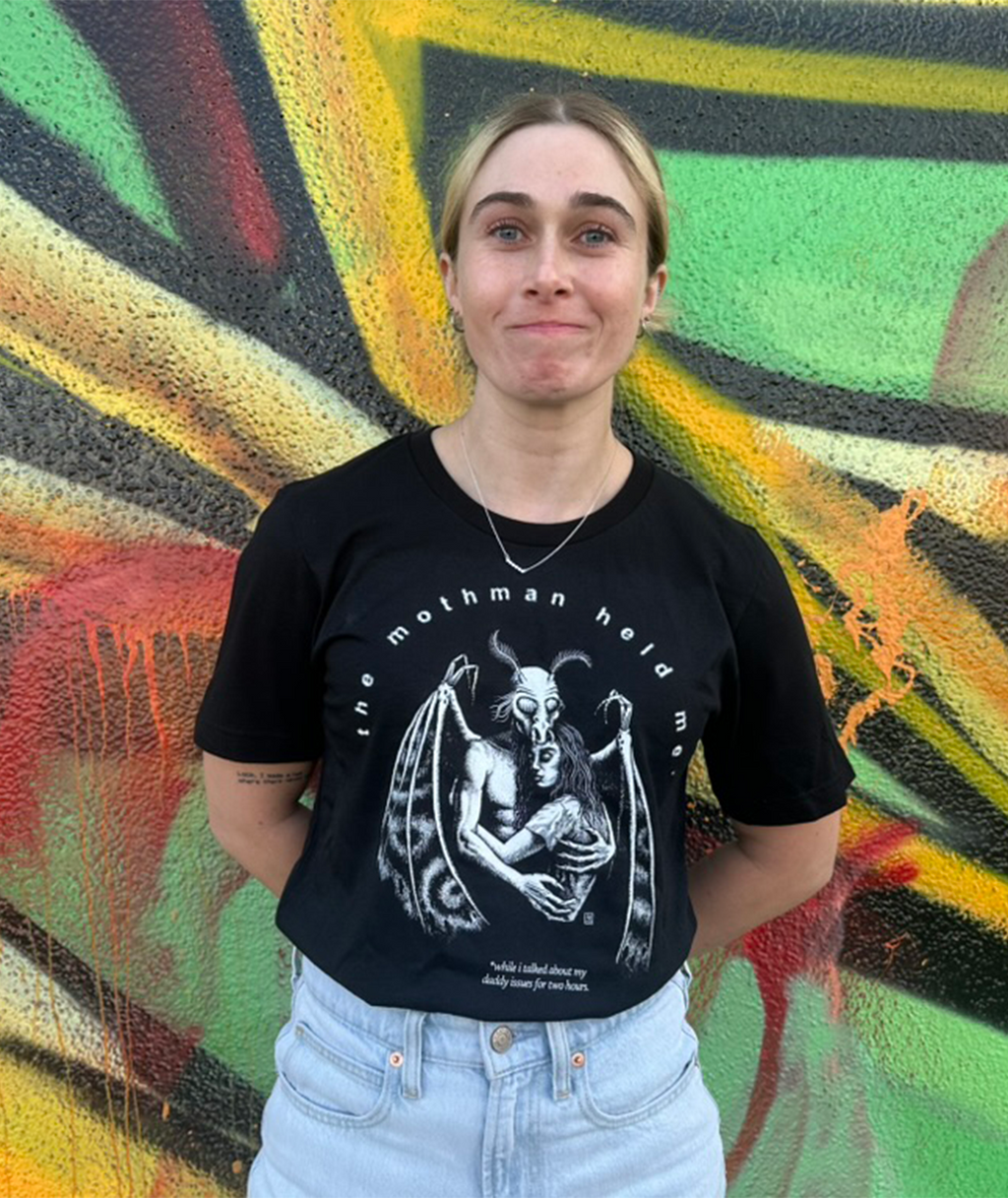 Beth May wearing a black t-shirt with a white design that says "the mothman hold me" with a mothman holding a woman. She is standing in front of a colorful, abstract wall.