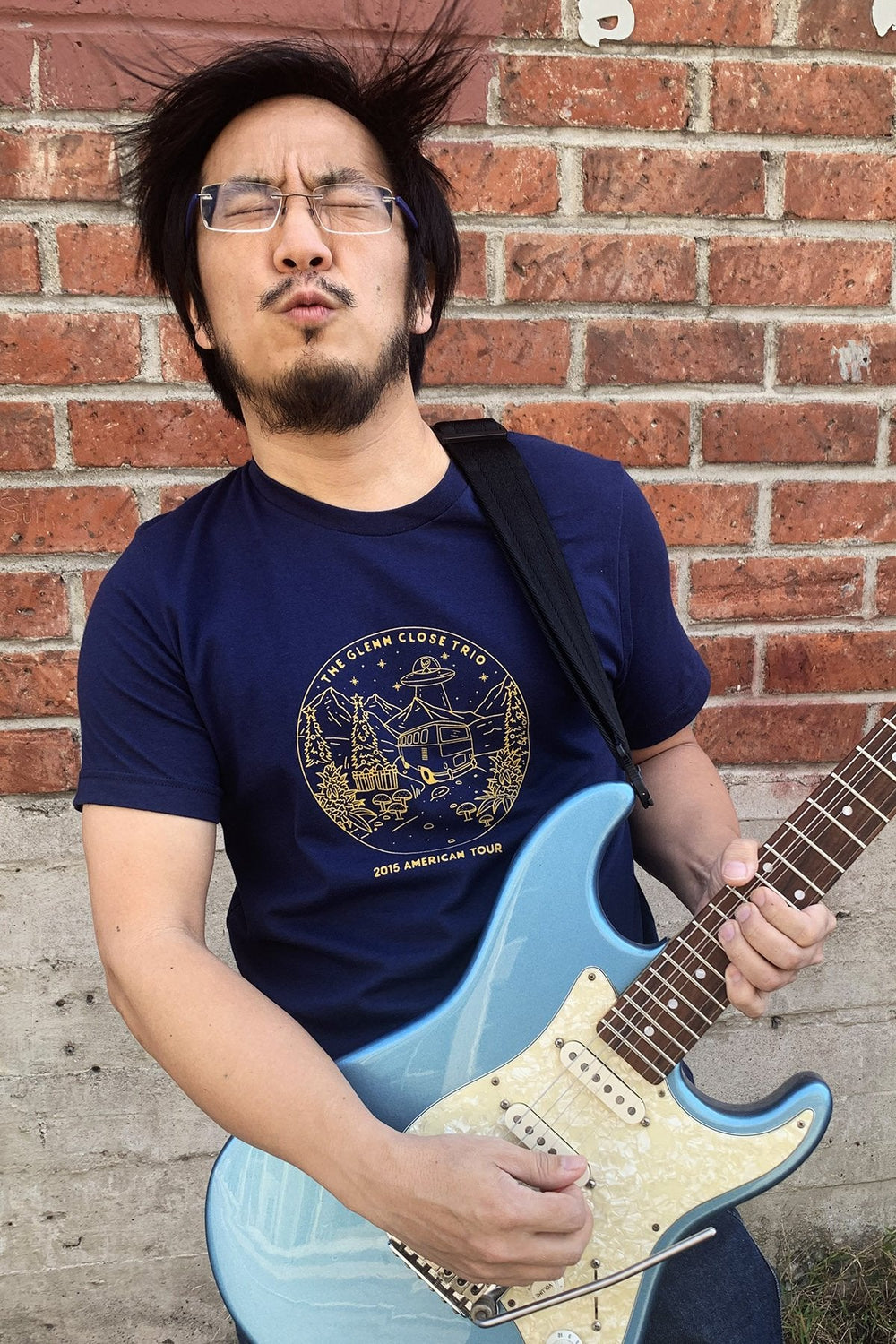 Freddie Wong wearing the t-shirt and playing a light blue electric guitar in front of a brick wall.
